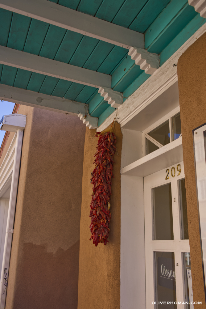 Brown Adobe building with white wood door with number 209 in metal numbers and chili ristra hanging from a blue and white overhang. Sante Fe, New Mexico